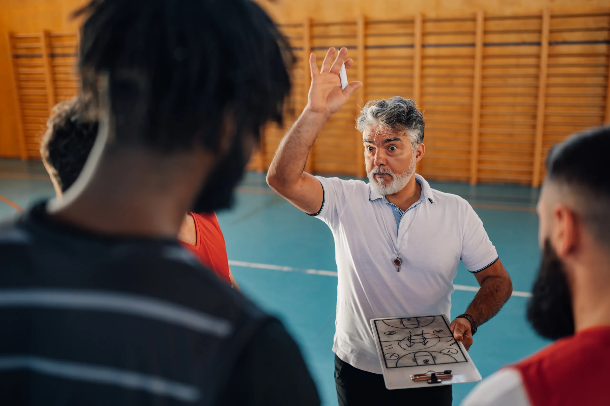 Basketball coach giving a team strategy session on the court, holding a clipboard with a play diagram.