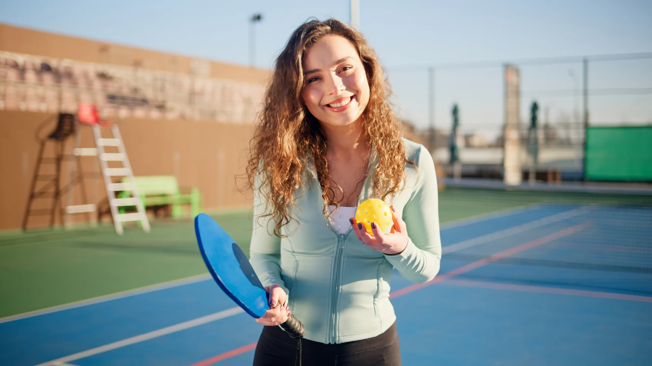 Smiling woman holding a pickleball paddle and ball, standing on a sports court.