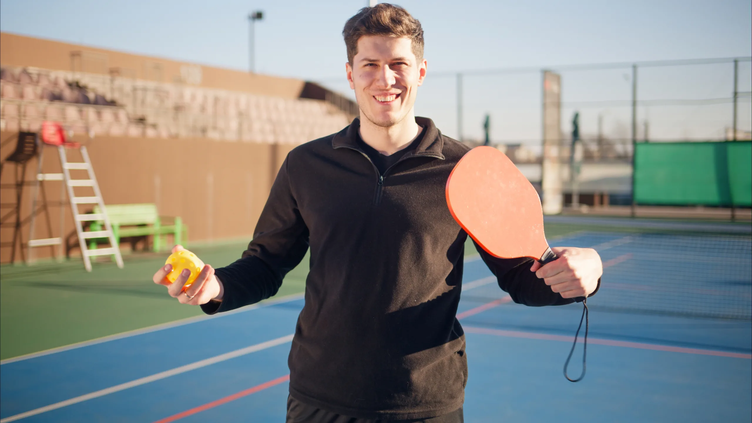 Smiling person holding a pickleball paddle and ball on an outdoor court under a clear sky.