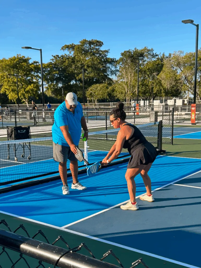 Man teaching woman pickleball technique on outdoor court. Sunny day, trees in background.