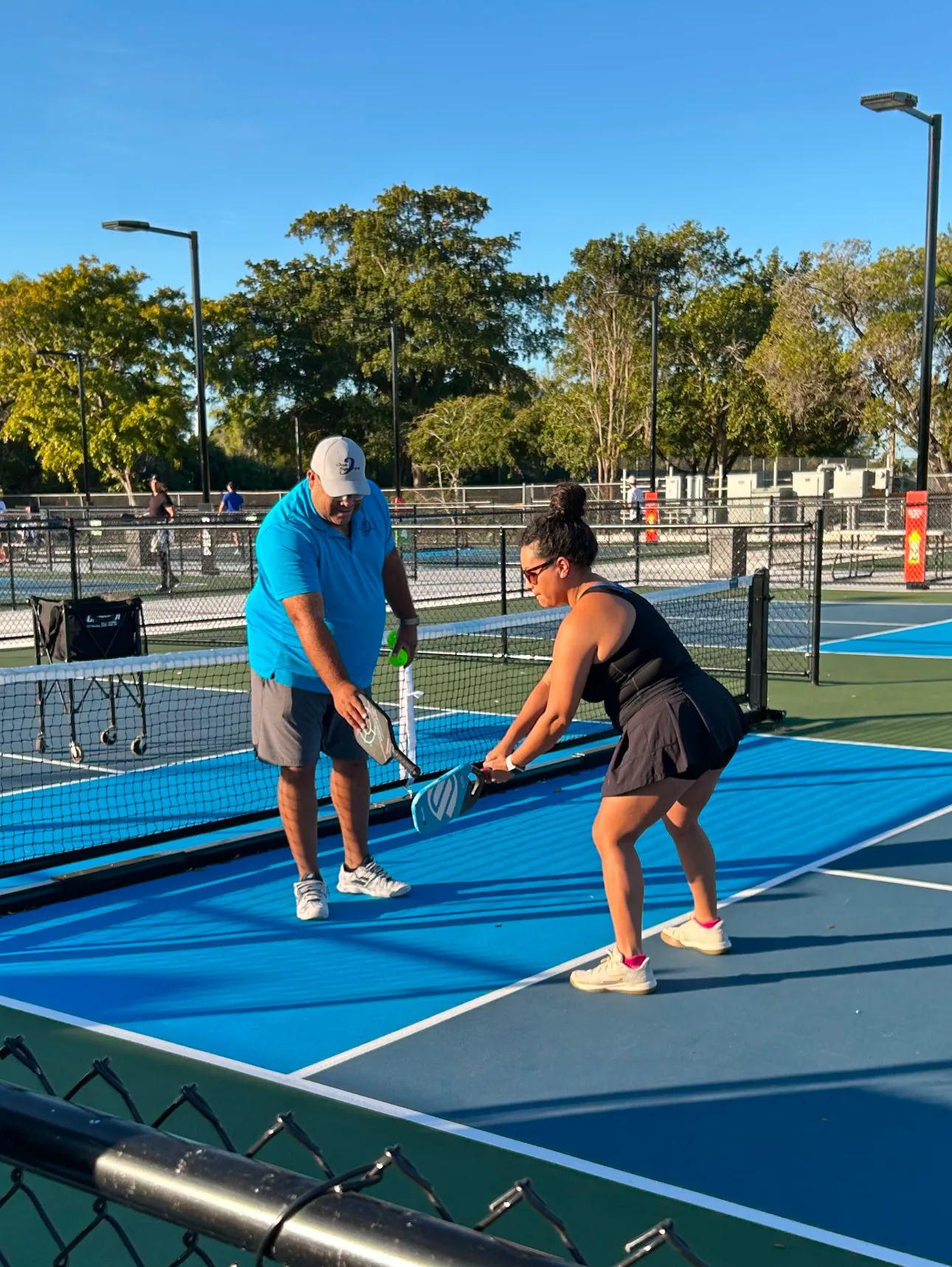 Man teaching woman pickleball technique on outdoor court. Sunny day, trees in background.