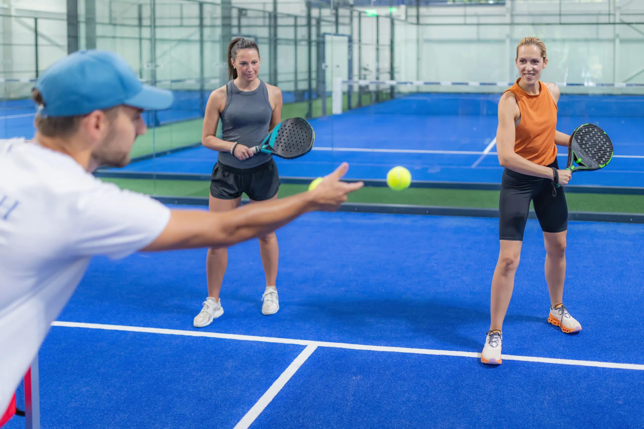 People playing padel on an indoor court; one is tossing a ball, and two others hold rackets, ready to hit.