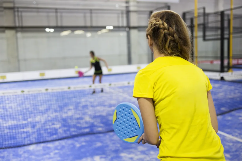 Two women playing padel on a blue court, one holding a blue racket, wearing a yellow shirt, with a net in between.