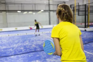Two women playing padel on a blue court, one holding a blue racket, wearing a yellow shirt, with a net in between.