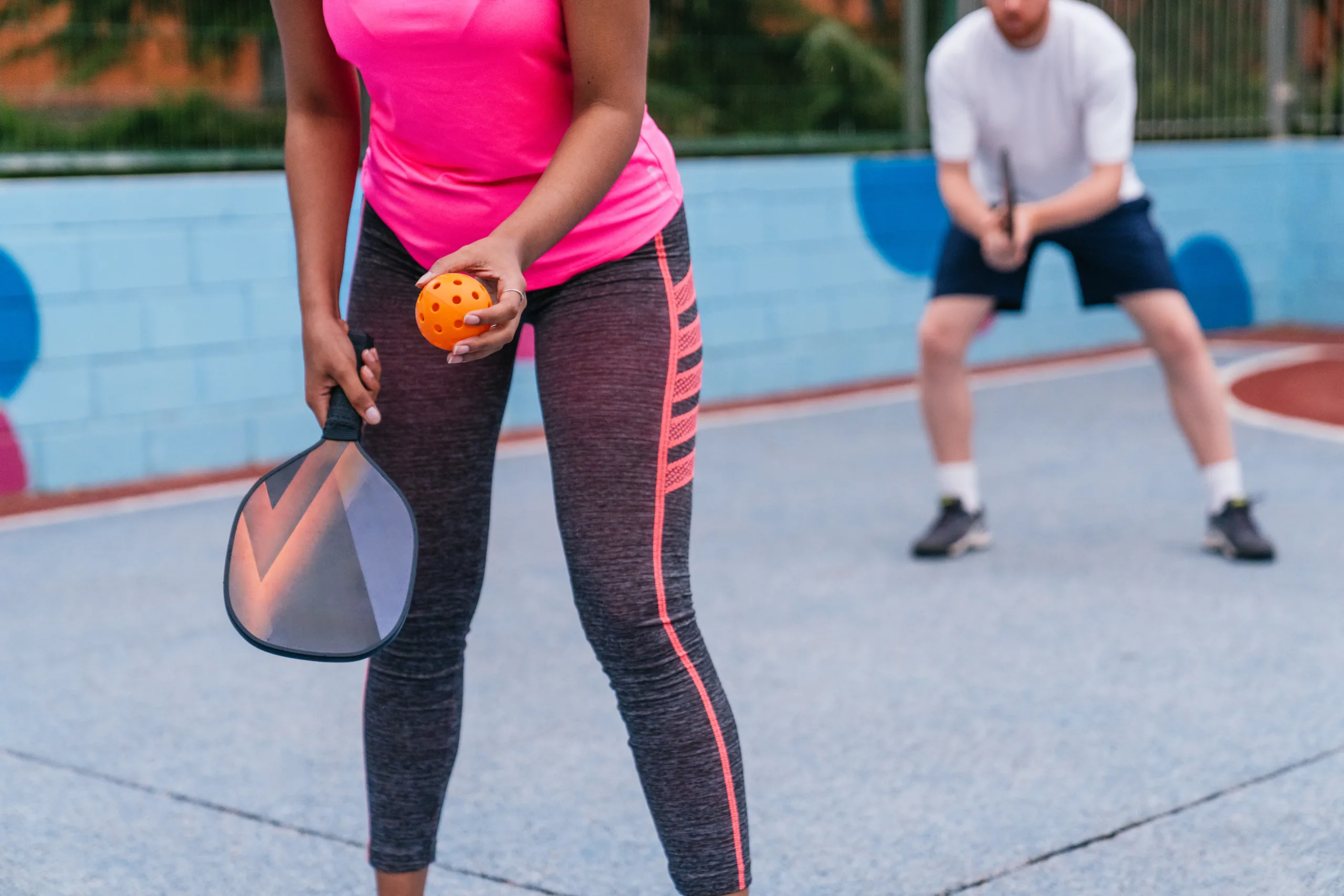 Two people playing pickleball, one about to serve with a paddle and orange ball on an outdoor court.