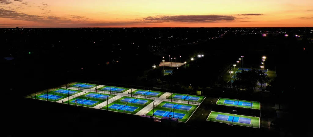 Lit tennis courts at dusk, with a vibrant orange and purple sunset in the background.